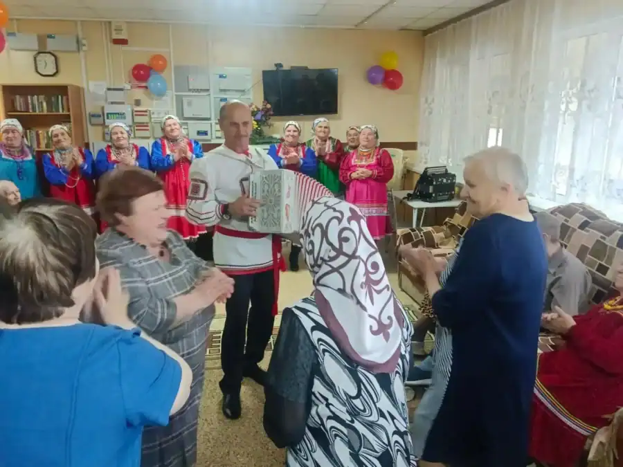 Мужчина играет на гармони среди женщин в народных костюмах и зрителей.
