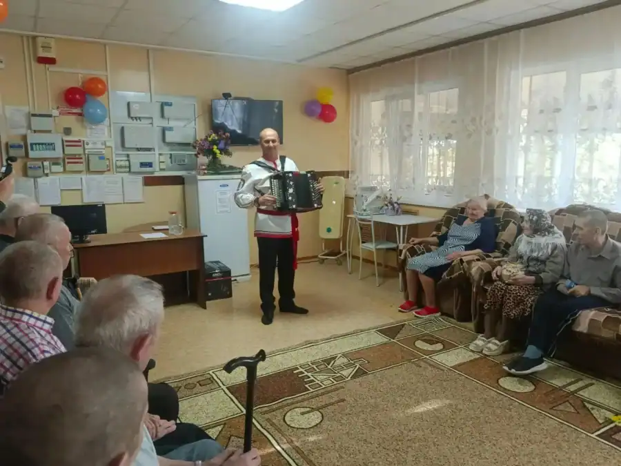 Музыкант с гармонью выступает перед сидящими пожилыми зрителями.