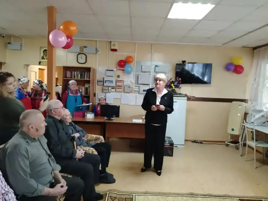 Женщина в деловом костюме выступает перед пожилыми зрителями.