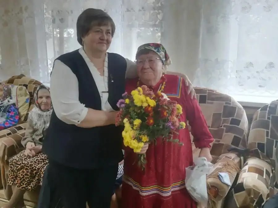 Женщина вручает пожилой участнице в народном костюме большой букет.