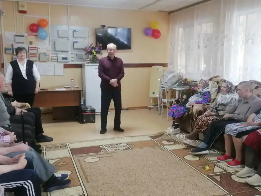 Мужчина стоит перед сидящими по обе стороны комнаты пожилыми людьми.