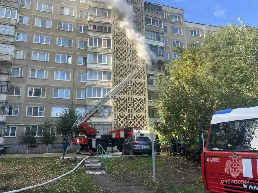Пожарная автолестница у многоэтажного дома, из верхней части которого идёт дым.