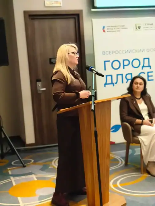 Женщина говорит у трибуны с микрофоном, рядом сидит участница сессии.