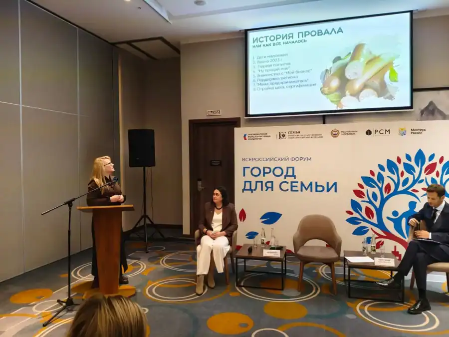 Выступающая стоит у трибуны перед стендом форума и экраном презентации.