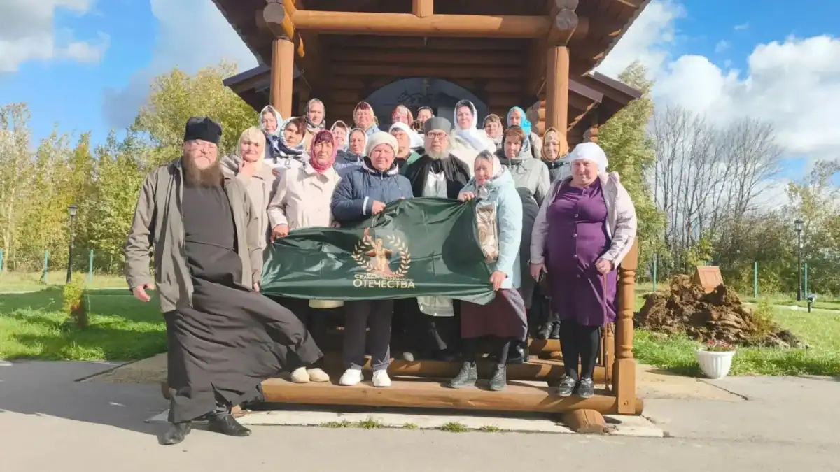 Группа людей у деревянной часовни держит зелёный флаг Комитета семей воинов Отечества.