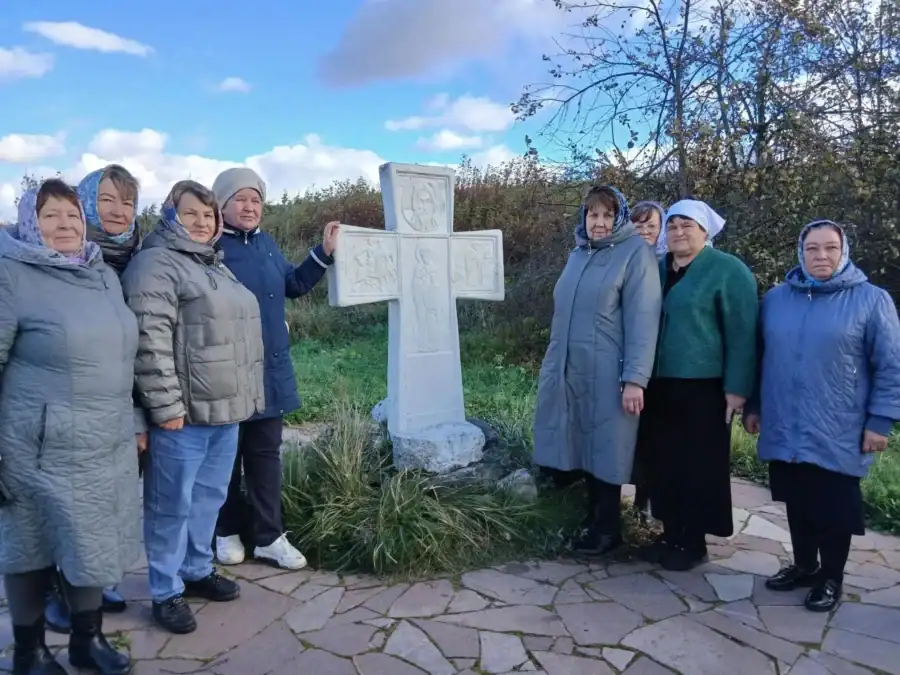 Женщины стоят рядом с белым резным поклонным крестом на природной территории.