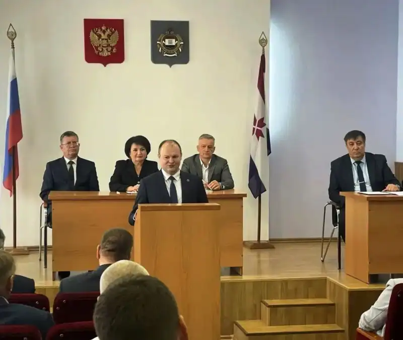 Мужчина выступает за трибуной в зале заседаний перед депутатами.
