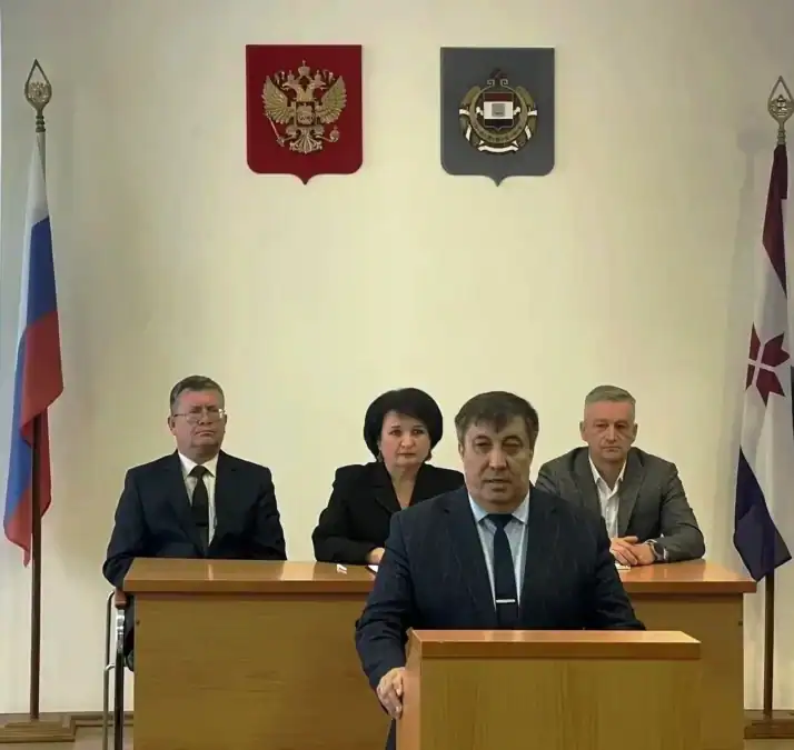 Мужчина выступает за трибуной, позади него сидят члены президиума.
