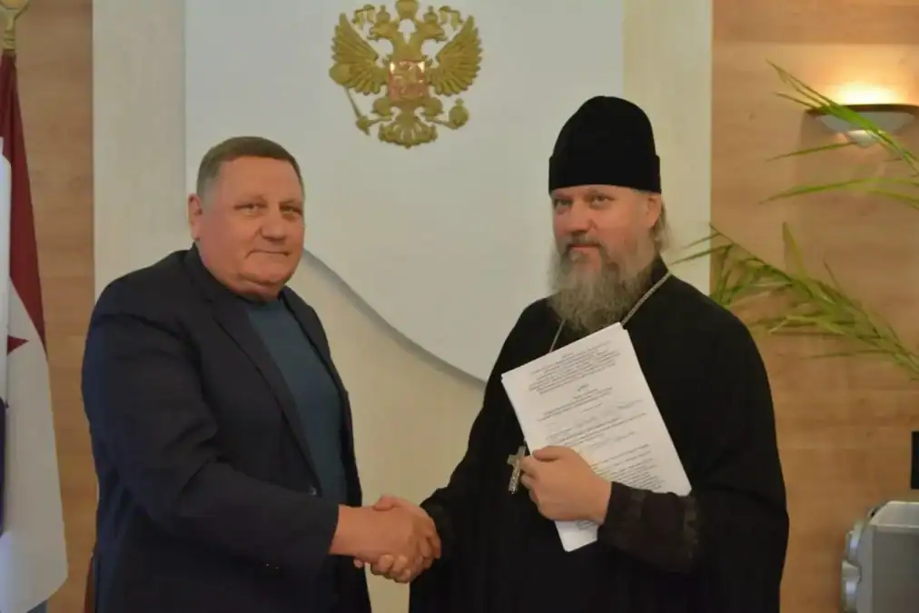Мужчина в церковном облачении пожимает руку представителю администрации, держа документ.