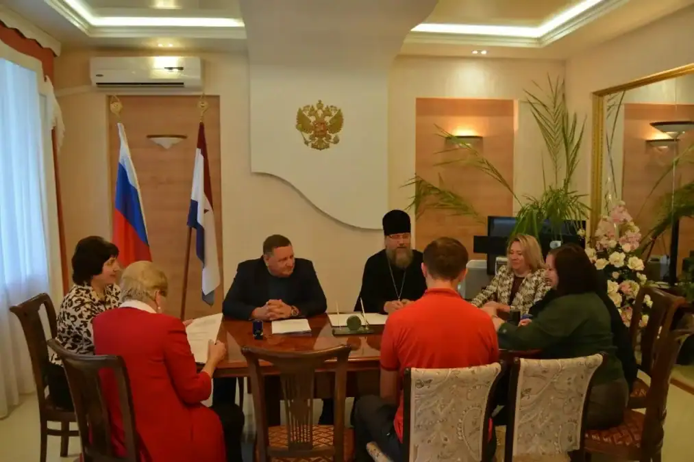 Участники встречи сидят за столом в зале с российским флагом.
