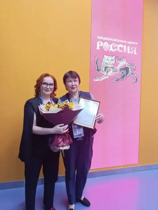Две женщины с бейджами держат букет и благодарственное письмо у стенда центра «Россия».