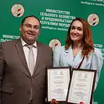 Женщина держит два диплома рядом с мужчиной у зелёного стенда.