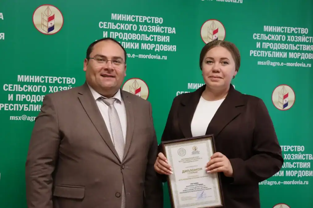 Женщина держит диплом рядом с мужчиной на фоне ведомственного стенда.