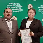Женщина держит диплом рядом с мужчиной на фоне ведомственного стенда.