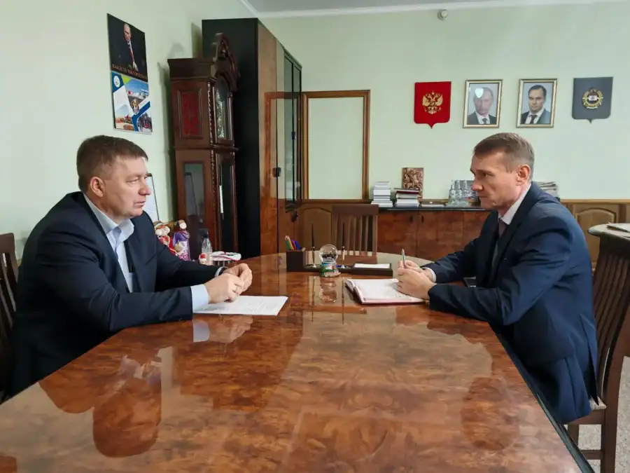 Двое мужчин в деловых костюмах беседуют за столом с документами в кабинете.