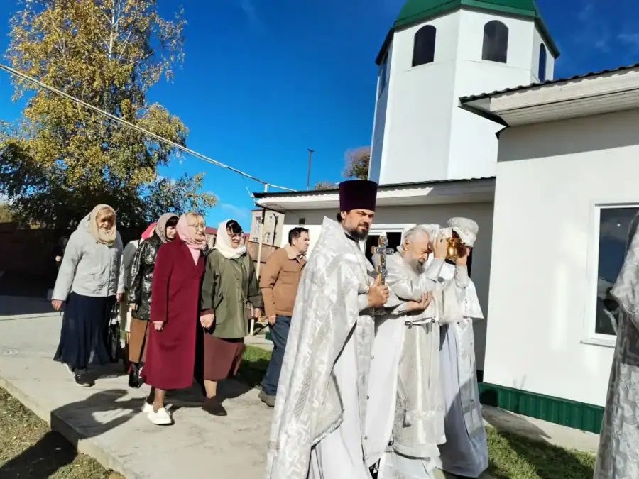 Священнослужители и прихожане идут процессией возле белого храма с колокольней.