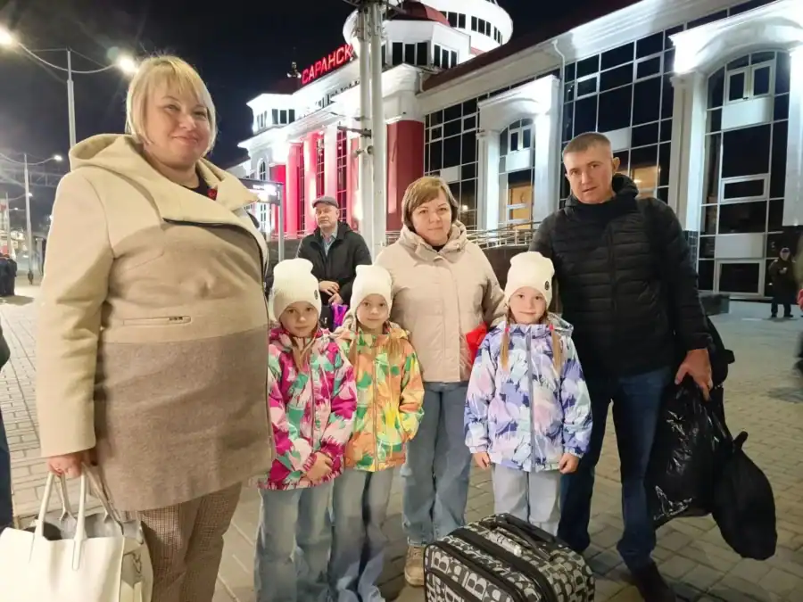 Семья с тремя девочками стоит с чемоданами у вокзала Саранска вечером.