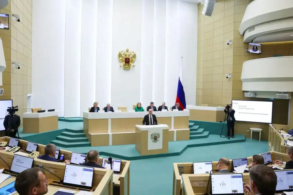 Докладчик выступает с трибуны в зале заседаний перед президиумом.