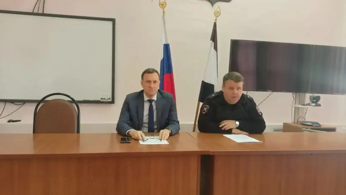 Двое мужчин сидят за столом заседания на фоне флагов и экранов.