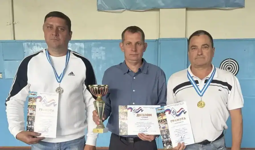 Трое мужчин позируют с кубком, дипломами и медалями у мишени.