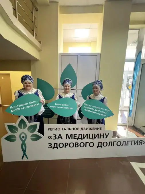 Три женщины в народных костюмах держат тематические таблички у стенда.