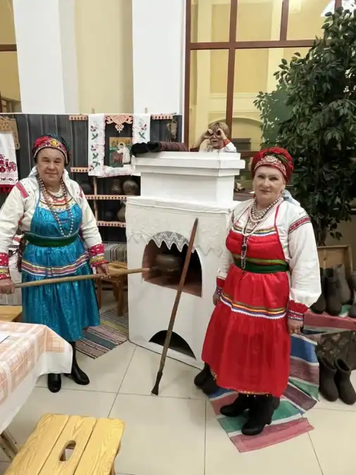 Женщины в народных костюмах стоят у белой печи и ремесленной экспозиции.