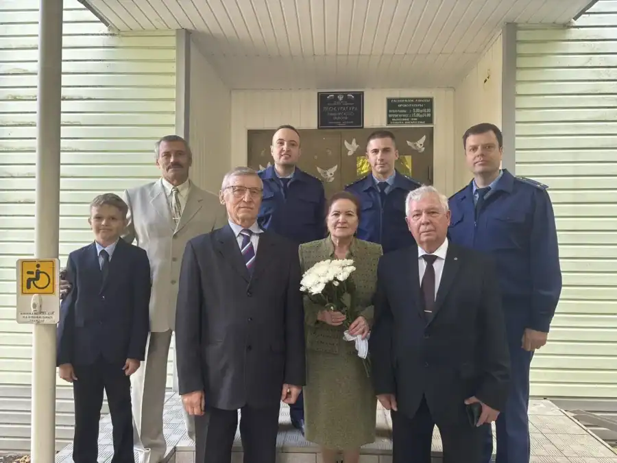 Группа людей у входа в здание, женщина в центре держит букет белых цветов.