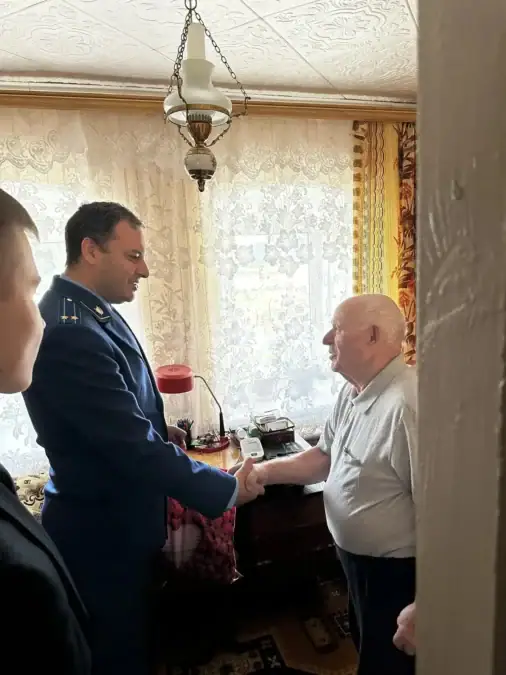 Сотрудник прокуратуры пожимает руку пожилому мужчине в жилой комнате.