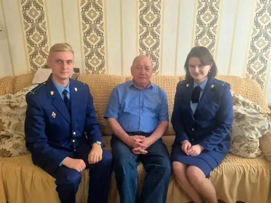 Пожилой мужчина сидит на диване между двумя сотрудниками прокуратуры.