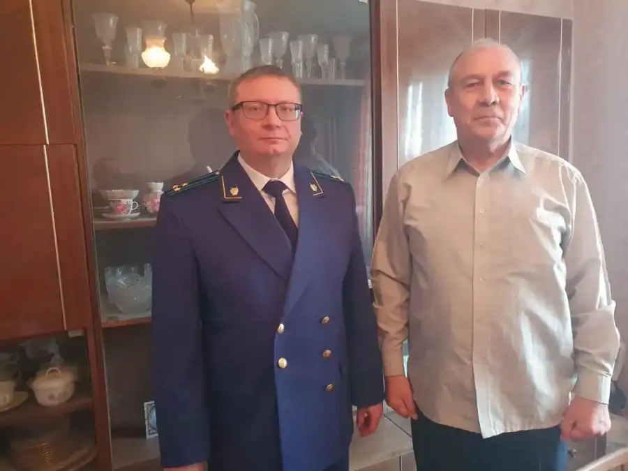 Сотрудник прокуратуры и пожилой мужчина стоят в комнате рядом с сервантом.