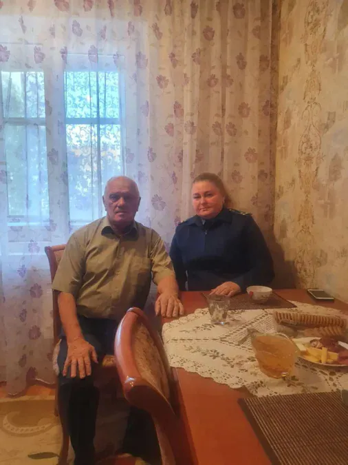 Пожилой мужчина и сотрудница прокуратуры сидят за накрытым столом.