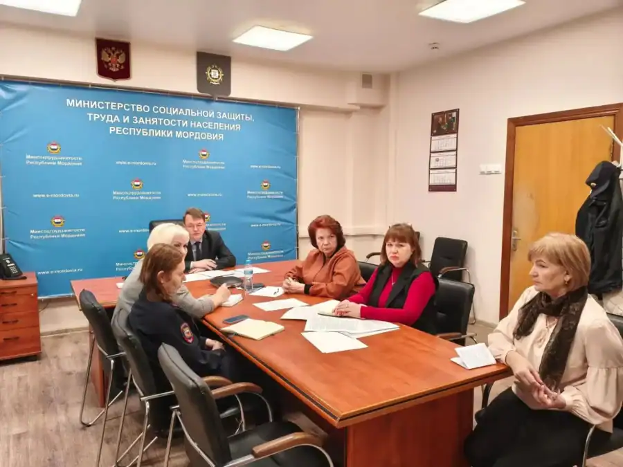 Участники заседания с документами сидят на фоне стенда министерства.