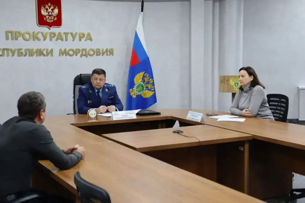 Прокурор и женщина принимают посетителя за столом в служебном кабинете.