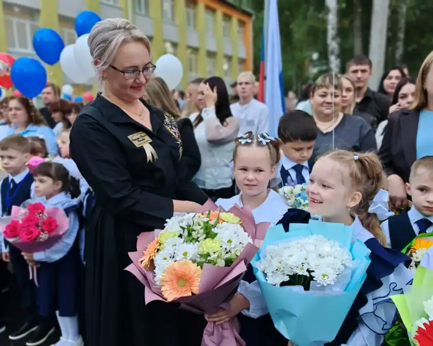 Учительница и младшие школьники с букетами стоят на праздничной линейке.