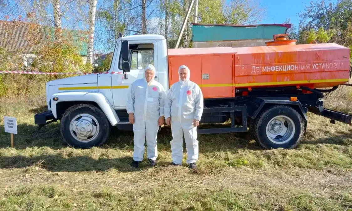 Два сотрудника в защитных костюмах стоят у дезинфекционной автоцистерны.