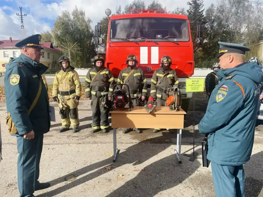 Пожарные в экипировке стоят у стола с инструментами перед пожарной машиной.