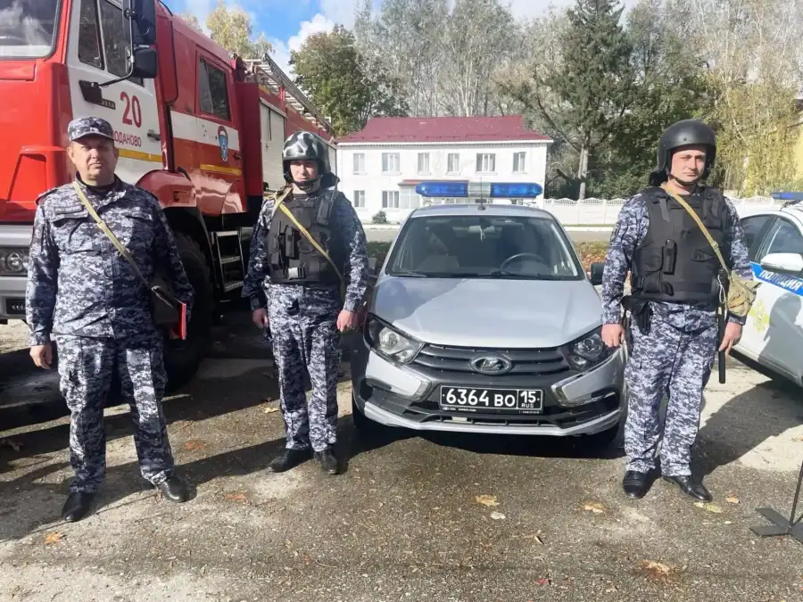 Трое сотрудников в защитном снаряжении стоят у пожарной машины и автомобиля.