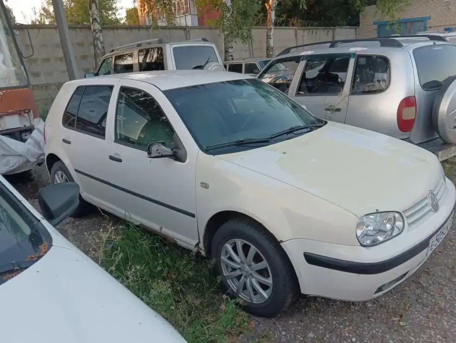 Белый Volkswagen Golf стоит среди других автомобилей во дворе.