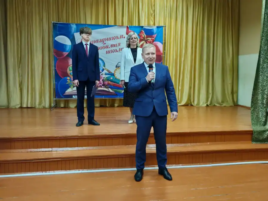 Мужчина с микрофоном выступает на сцене перед юбилейным баннером школы.