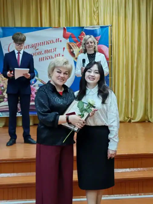 Две женщины на сцене держат букет роз, позади стоят ведущие.