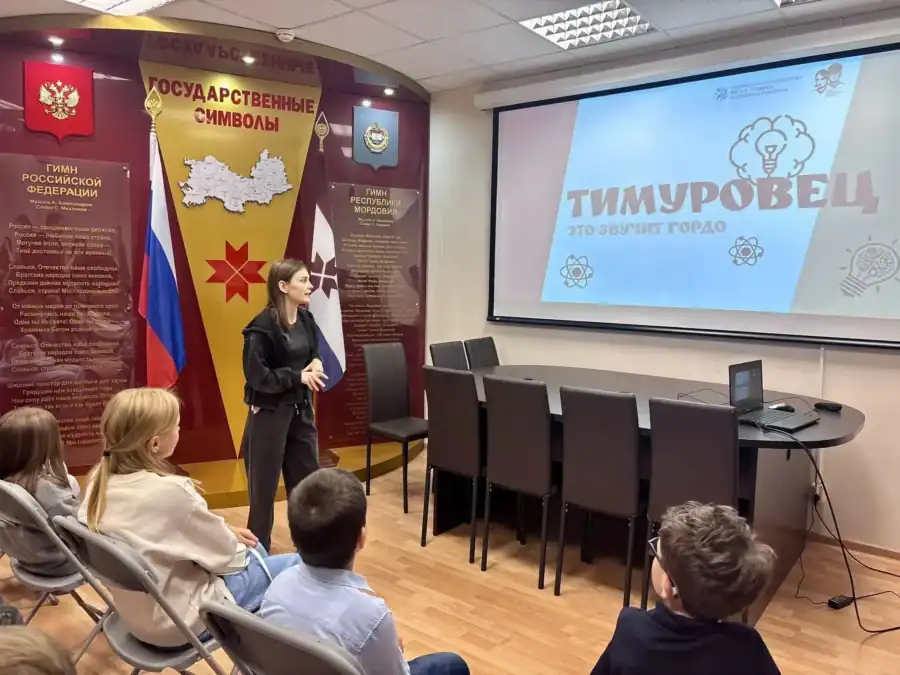 Сотрудница выступает перед детьми у экрана с презентацией о тимуровцах.