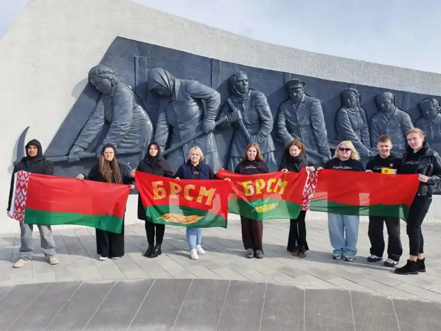 Группа молодых людей с белорусскими флагами стоит у монументального рельефа.