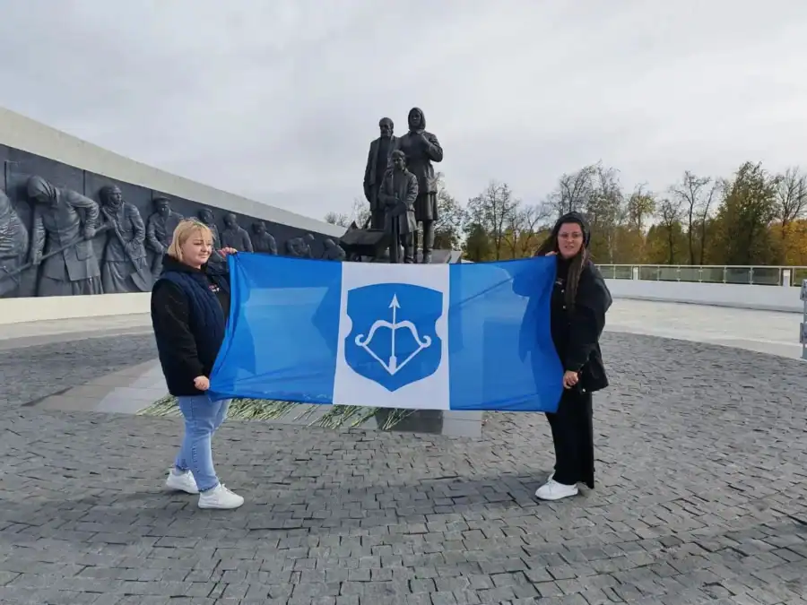 Две женщины держат синий флаг перед мемориальным комплексом со скульптурами.