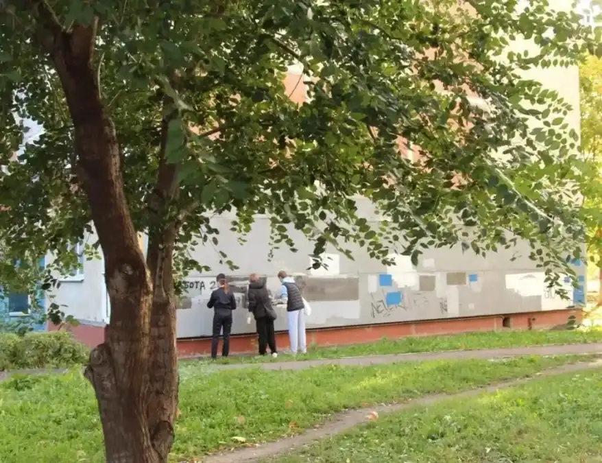 Трое человек стоят у фасада жилого дома под большим деревом.