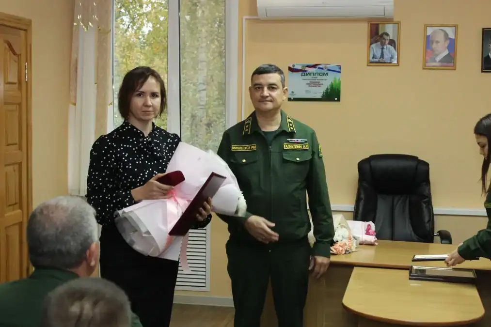 Женщина держит букет и документы рядом с сотрудником лесной службы в кабинете.