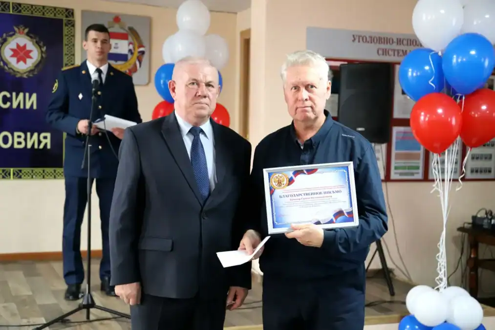 Двое мужчин стоят в зале, один держит грамоту и конверт.