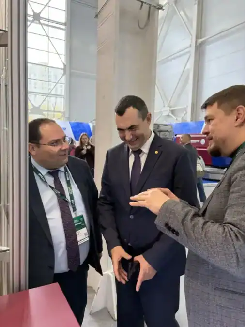 Трое мужчин беседуют у выставочного стенда, один показывает жестом на экспозицию.