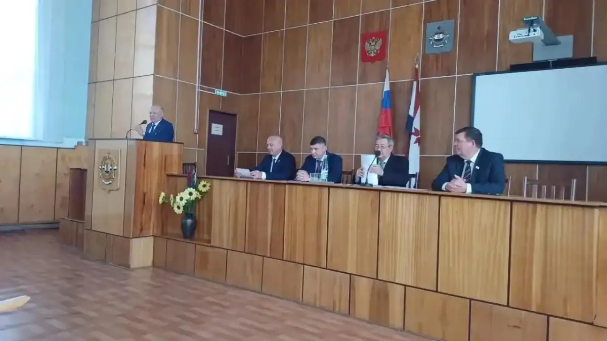 Выступающий стоит за трибуной перед президиумом в зале заседаний.