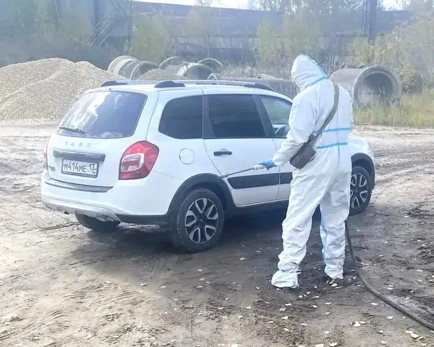 Специалист в защитном костюме обрабатывает белый легковой автомобиль.