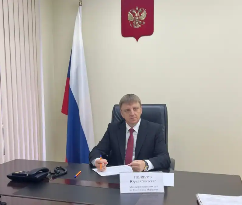 Мужчина в костюме сидит за столом с документами, российским флагом и гербом.